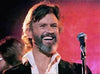 A bearded man with wavy hair smiles and stands in front of a microphone on stage, illuminated by pink and purple lights, capturing the laid-back charm reminiscent of Willie Nelson's performance as seen on the "Songwriter (1984) DVD.