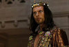 A man, adorned in an ornate crown and jeweled robe, stares intensely, embodying the grandeur of *One Night with the King (2006)*. With long dark hair and posed before a backdrop of blurred architectural details, he captures the epic and historical drama brought to life in this DVD.
