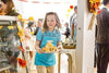 A young girl in a blue uniform carries a plate of pastries at a festive indoor gathering adorned with autumn leaves, while guests mingle in the background—capturing the heartwarming, small-town bakery charm found in “Truly Madly Sweetly (2018)” DVD.