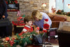 A woman and a young boy decorate a desk with Christmas ornaments and garland in a festively adorned office with stone walls, surrounded by holiday decorations and the warm glow of “A Gift Wrapped Christmas (2015) DVD” playing quietly in the background.