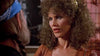 A woman with curly brown hair and hoop earrings talks to a man in a blue bandana. The dim, gently blurred background suggests the laid-back ambiance of a bar or club, where something like Songwriter (1984) on DVD might be playing quietly in the corner, adding to the easygoing vibe.