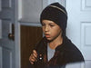 A young boy wearing a dark knit hat and jacket stands indoors, gazing downward with a serious look. He clutches the strap of his backpack, appearing ready for an adventure—or perhaps preparing to settle in and watch “When Angels Come To Town (2004)” on DVD. In the background, a door and wall are visible.
