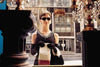 A woman in a black dress, pearl necklace, tiara, and sunglasses stands outside a store window holding a white bag with ‘Schodts’ on it—a scene evocative of “The Audrey Hepburn Story (2000) DVD.” Chandeliers and a city street appear in the background.