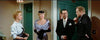 Four well-dressed people, two women and two men, converse in a hallway by green doors, creating the suspenseful atmosphere of a Hollywood whodunit. One woman wears a purple dress and gloves; the other sports a light sweater. The men are dressed in suits. An exit sign hangs above one door—an ambiance evocative of classic movies like Black Widow (1954), now available on DVD for fans of rare, hard-to-find films from cinema’s golden age.
