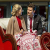 A man in a suit and a woman in a red dress with a beige shawl sit closely together on a decorative sleigh, sharing a festive blanket. Snowflake decorations and holiday lights twinkle in the background, setting the stage for classic holiday romance just like that found in With Love, Christmas (2017) DVD.