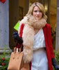 A woman with blonde hair, dressed in a red coat, white dress, large furry scarf, and black gloves while holding a tan handbag near a building entrance, evokes the elegance and charm reminiscent of “A Gift Wrapped Christmas (2015)”—now available on DVD.