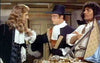 Three men in period costumes stand indoors: one like a pirate with a blonde wig, another in a black hat and white collar holding a book, and the third posing by gold objects—evoking scenes from "Morgan The Pirate" DVD (1960).