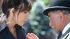 A woman with dark hair faces an older man in a bowler hat and suit, engaging in a serious conversation outdoors—a scene reminiscent of the film “When Angels Come To Town (2004)” on DVD.