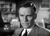 Black-and-white photo of a man in a suit, looking serious, with short hair. The background is filled with blurred shelves and a framed picture on the wall—evoking the timeless atmosphere captured in classic films like *Dodsworth (1936) DVD*, reminiscent of the era when directors such as William Wyler shaped cinematic history.