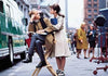 A man sits on a director’s chair on a city street film set, while a woman in a trench coat—reminiscent of Jennifer Love Hewitt’s role in *The Audrey Hepburn Story (2000) DVD*—stands beside him holding books. Film equipment and crew members are visible in the background, evoking the timeless appeal found in classic movie collections.