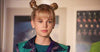 A young person with blonde hair styled in two space buns, silver hoop earrings, and a vibrant jacket stands indoors against a backdrop of blurred framed photos—evoking the futuristic style of Zenon from “Zenon: Girl of the 21st Century (1999) DVD.”.