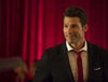 A man in a dark suit and red tie stands in front of red curtains, smiling slightly—as if he’s just stepped out of “With Love, Christmas (2017) DVD.” The softly blurred background creates the warm, inviting atmosphere you’d expect from this timeless holiday movie.