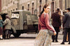 A young woman with braided hair and a long skirt looks over her shoulder on a bustling street, where passersby and an old military vehicle marked with a swastika recall evocative scenes from “The Audrey Hepburn Story (2000) DVD,” reminiscent of those found in rare classic movie collections or biographical films about the iconic actress.