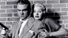 A serious man in a suit holds a gun while a concerned woman clings to him from behind, standing before a brick wall in a dramatic black-and-white scene that captures the suspenseful atmosphere of classic noir crime thrillers—perfectly evoking the style of rare, hard-to-find films like “Kiss Tomorrow Goodbye (1950)” now available on DVD for collectors.