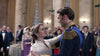 A woman in a white dress and a man in a royal blue uniform dance together at a formal event, surrounded by elegantly dressed guests—a scene evoking the enchanting atmosphere of *A Christmas Prince: The Royal Wedding (2018)*, now available on DVD.