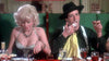 A woman in a sparkly dress and a man in a black hat and suspenders are eating at a table, evoking scenes from the “Death Race 2000 (1975) DVD”; the man is messily licking his fingers with a napkin tucked into his shirt.