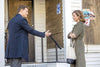 A man in a suit gestures with his hand while talking to a woman in a trench coat standing on a porch with boarded windows and campaign posters, creating a scene reminiscent of those found in offbeat romantic comedies like the “Truly Madly Sweetly (2018) DVD.”.