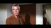 A serious-looking man in a brown suit stands indoors, his reflection visible in a nearby mirror—evoking the moody atmosphere of the neo-noir thriller “Point Blank (1967) DVD”—as he is positioned near a closed wooden door, with light and shadows cast on the wall behind him.
