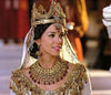 A woman adorned in an ornate gold crown, jeweled headpiece, and elaborate gold jewelry over a white dress gazes off to the side with a smile, evoking the grandeur of a biblical epic. The softly blurred background enhances the timeless atmosphere, reminiscent of a scene from the “One Night with the King (2006) DVD.”.