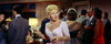 A woman in a pink dress holds a cigarette holder at a lively indoor party, capturing the glamorous intrigue of classic Hollywood as seen in Black Widow (1954). Elegantly dressed guests mingle in the background, evoking scenes reminiscent of rare films like this hard-to-find DVD.