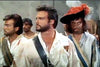 Three men in period costumes pose together; the central, bearded figure in a torn white shirt and blue sash recalls Steve Reeves in a classic pirate film. On the right, a man wears a black hat with an orange feather. Product: Morgan The Pirate DVD (1960).