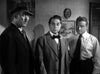 Three men in formal attire stand indoors; one wears a hat and suit, another has a bow tie and vest, and the third is in a shirt and tie—a scene evocative of classic films like “Kiss Tomorrow Goodbye (1950)” on DVD. A framed picture and a round clock are visible on the wall behind them.