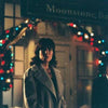A woman in a light coat stands outside at night near a festive archway adorned with colorful holiday lights, evoking the heartwarming atmosphere of *When Angels Come To Town (2004)*, with a sign reading Moonstone Bar illuminated in the background.