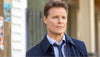 A man with short, light brown hair wearing a blue dress shirt, tie, and dark coat stands outdoors, looking thoughtful—evoking the leading character from the charming small-town bakery movie “Truly Madly Sweetly (2018) DVD.”.