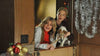 Two women smile and look outside while holding a small dog. Holiday decorations, including Christmas ornaments and silver tinsel, surround the doorway, creating a heartwarming scene reminiscent of a family moment from the “Holiday Road Trip (2013) DVD.”.
