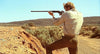 A person in a tan shirt and olive pants aims a rifle over a rocky, shrub-dotted landscape beneath a clear blue sky, reminiscent of the vivid outback scenes featured in the Wake in Fright (1971) DVD.