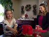 Two women sit at a festively decorated table. One woman carefully folds a scarf while the other, holding a cheerful mug, smiles warmly. Holiday wreaths, garlands, and Christmas decorations fill the background—evoking the cozy scenes captured in “A Gift Wrapped Christmas (2015) DVD.”.