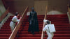 Certainly! Here’s a rewritten version of the sentence incorporating the product data for “Death Race 2000 (1975) DVD”: A person in a black cape and mask stands on red-carpeted stairs, while two people in white suits—possibly collectors searching for rare movies like Death Race 2000 (1975) on DVD—face them from either side of the stairway, one holding a large camera.