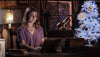 A woman sits at a desk using a laptop in a cozy room with a glowing lamp and a decorated white Christmas tree behind her, perhaps searching for “A Christmas Prince: The Royal Wedding (2018) DVD” or reminiscing about favorite holiday films to enjoy during the festive season.
