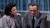 A woman with dark hair and a decorative blouse sits beside a man in a gray suit and tie, engaged in a tense conversation before blue curtains—an atmosphere evocative of classic Hollywood mysteries like the 1954 film “Black Widow,” now available on DVD.