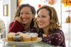 Two women smile and lean on a glass counter with a plate of colorful cupcakes in front of them, evoking the warm charm of a small-town bakery—just like a scene from “Truly Madly Sweetly (2018)” on DVD. The bright, fall-themed décor in the background creates a cozy atmosphere perfect for this romantic comedy.