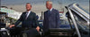 Two men in suits stand beside a sleek black convertible under the bright sun, capturing the sharp, stylish aesthetic of a neo-noir thriller. One man grins while holding open the car door; the other stands close, glancing at his companion. In the background, cars, power lines, and street signs set a scene evocative of classic films like Point Blank (1967)—now available on DVD.