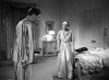 A man in striped pajamas stands facing a woman in a gown and headwrap in a vintage bedroom, evoking the timeless elegance of classic films like “Dodsworth (1936)” on DVD. The woman appears upset, standing near a bed draped with a satin comforter. The room is elegantly furnished and softly lit.