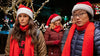 Three people outdoors at night wear Santa hats, red scarves, and winter jackets. Holiday lights and trees twinkle in the background. Two stand seriously in front while a third lingers just behind—resembling a scene straight from the Love Hard (2021) DVD.