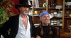 Two men stand side by side in a room filled with shelves of books and tapes, alongside a collection that includes the "Songwriter (1984) DVD." One wears a black hat and jacket over an open shirt, while the other sports a blue bandana, braids, and a black shirt—both smiling warmly.
