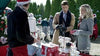 A man and woman shop at an outdoor Christmas market, browsing festive items on a table, including the “With Love, Christmas (2017)” DVD—an ideal holiday romance for cozy nights. The vendor sports a Santa hat and red apron, while evergreen trees and holiday decorations create a festive backdrop.