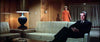 A man in a suit sits on a beige couch in a modern, wood-paneled living room, capturing the stylish atmosphere of the Point Blank (1967) DVD. In the background, a woman in an orange dress stands near drawn curtains, infusing the scene with an air of intrigue reminiscent of this neo-noir classic.