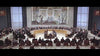A large group of people sit in a circular arrangement at a formal meeting in a grand room with tall windows and an impressive mural, evoking scenes from classic disaster films like “Meteor (1979)” featuring stars such as Sean Connery and Natalie Wood.