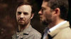 A man with a serious expression looks at another man in glasses and a beige coat in a dimly lit setting, enhancing the mystery of We Go On (2016) DVD, a rare film about life after death.
