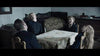Four people sit around a table in a dimly lit, old-fashioned room, appearing lifeless. The eerie stillness mirrors the chilling atmosphere found in the supernatural horror film "Post Mortem (2020)" DVD.