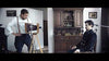 A man uses an old camera on a tripod to photograph another man in a dark jacket inside a vintage, wood-furnished room—evoking scenes from the Post Mortem (2020) DVD, with framed pictures and a large cabinet on the walls.