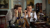 Two men sit at a cluttered desk talking on black phones. One wears a white naval uniform with epaulets—a scene reminiscent of a World War II drama like Pursuit of Graf Spee (1956) DVD. Candles, statues, and framed photos decorate the room.