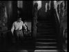 A man in glasses and a white shirt stands by a staircase in eerie lighting, evoking the suspense and vintage horror style of classic films like The Ghoul (1933) DVD.