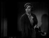 Black and white image of a woman in a hat, scarf, and coat stands indoors with a serious look—capturing the moody intrigue of vintage horror like "The Ghoul (1933) DVD." Shadows fall across minimal background details.