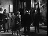 Five people stand in a dimly lit, ornate room with stone walls and a staircase—echoing vintage horror cinema—in this atmospheric black-and-white scene from The Ghoul (1933) DVD. Three face forward while two, center and right, have their backs turned.