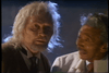 Two older men stand close together; one, with long white hair and a beard, is dressed in a suit, while the other has short gray hair, a mustache, and wears a white lab coat. They seem to be engaged in a serious discussion—perhaps debating the kind of time-travel exploits found in classic cyborg action films like *Eliminators* (1986), now available on DVD.